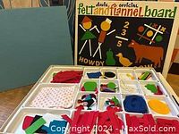 Felt and flannel board with box and colorful felt shapes inside white plastic tray compartments.