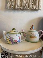Two vintage ceramic tea pots displayed side by side on a round table. One tea pot decorated with multicolor flowers and green accents, the other Peter Rabbit themed with rabbit figurine lid.