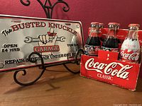 Six unopened Coca-Cola glass bottles with Dale Earnhardt NASCAR labels in Coca-Cola cardboard carrier beside one metal decorative automotive-themed license plate on display stand