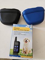 Remote dog training collar box with image of dog, display of remote with buttons, and two silicone treat pouches (black and blue) placed on either side.