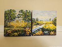 Both oil paintings shown side by side on floor against wall, displaying wooded and river landscape scenes with thick palette painting style strokes.
