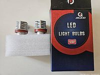 Pair of AOLEAD LED headlight bulbs with red mounting gaskets placed on white foam next to AOLEAD box