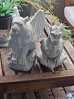 Photo of two medium-sized gray concrete/plaster statues, a gargoyle with wings on the left and a griffin with a horse head on the right, sitting on a wooden table with plants and outdoor furniture in the background.