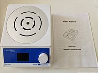 Front view of the ANZESER MS2000 magnetic stirrer showing the circular metal stirring surface, LCD screen, speed knob, and user manual beside it.