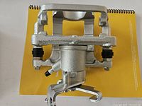 Photo showing one disc brake caliper assembly placed on a yellow notebook, viewed from the front and top to display general shape and mounting bracket layout.