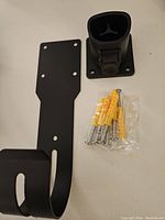 Photo of black stainless steel wall mount bracket with hook, black plastic nozzle holster, and mounting screws with yellow plastic anchors on a white surface.