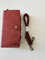 Closed red wallet case with button snap closure and detachable shoulder strap placed separately