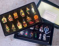 Photo showing two black trays with 24 assorted hand-painted European style blown glass Christmas ornaments in a green velvet box with a certificate of authenticity.