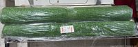 Two rolled artificial turf rugs on a wire rack, wrapped in plastic, labeled with lot number.