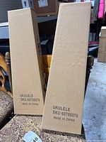 Two rectangular brown cardboard boxes upright on a chair, labeled UKULELE SKU:60700070 Made in China.