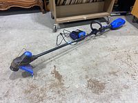Full length view of the blue and black Kobalt weed wacker with cutting head, handle, and battery compartment visible laid on concrete floor.