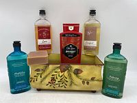 Photo showing grouping of five body wash bottles and two bar soaps placed inside a decorative metal tray with a leaf and berry pattern.