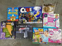 Overview photo showing all 8 unopened children’s games/toys including Headbanz 2, Clue, Laser X, Hot Tub High Dive, Mini Brands, Sequin Surprise llama craft kit, green blaster toy, and gold spinning top toy.