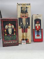 Front view of three nutcracker items in their original boxes: a large blue hat nutcracker, a medium red/blue holding sword nutcracker, and a ceramic cookie jar nutcracker with black hat.