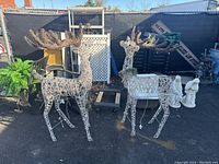 Pair of two large light-up reindeer decorations shown outdoors with black fencing and other miscellaneous items in background. Both reindeer are upright with wired white bodies and brown antlers, decorated with greenery.
