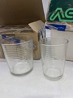 Two clear glasses with horizontal ribbed rim pattern, one taller glass and one shorter glass, placed in front of partially open cardboard boxes.