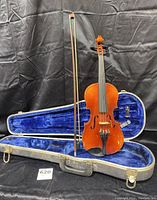 Full view of violin standing in open grey case showing blue plush interior, with bow placed inside case next to violin