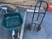 Metal wheelbarrow and handtruck side by side on parking lot pavement with orange cones in background