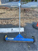 Full view of the blue magnetic sweeper with handle and two yellow wheels on asphalt surface.