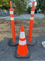 Photo showing three tall orange traffic cones with two white reflective bands each and weighted black rubber bases alongside one shorter cone.