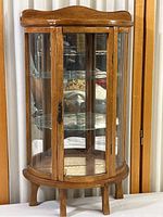 Front angled view of the vintage oak curio display cabinet showing rounded glass front door and sides, oak wooden top, and five short legs.