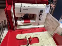 Beige and red Bernina sewing machine inside red plastic carrying case, accompanied by paperwork.