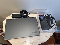 Closed gray Lenovo Yoga laptop alongside power adapter and clear bin of drives and peripherals