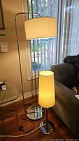 Full view of standing floor lamp with arched stainless steel base and rectangular fabric shade and table lamp with cylindrical fabric shade on wood floor near window and sofa