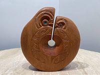 Front view of round wood sculpture showing central hole, koru patterns and two stylized heads with blue inlaid eyes