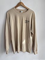 Front view of beige sweatshirt on hanger showing small black text on left chest and ribbed cuffs