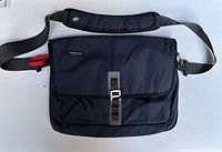 Dark navy Timbuk2 messenger bag showing front flap, adjustable padded shoulder strap with a red release lever and silver buckle.