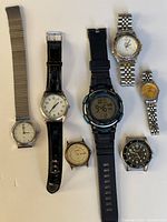 Photo showing six vintage watches of various styles and sizes arranged on a light background. Watches include different bands: metal expansion, leather strap, resin digital sports band, and linked metal bracelets.
