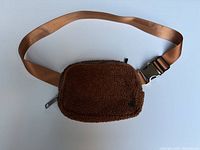 Front view of caramel-colored fleece belt bag with zipper and adjustable strap laid flat on white background.