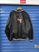 Back view of black Snap-On satin jacket with large embroidered wrench and Snap-On logo design, striped cuffs and waistband visible.