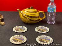 Photo showing the yellow and gold antique teapot next to three souvenir pads on a dark surface with a water bottle for scale.