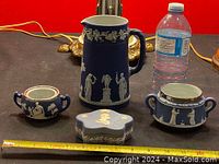 Photo of tall blue Wedgwood jasperware pitcher, two covered sugar bowls, oval lidded box, and water bottle for scale on black surface.