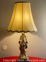 The lamp lit, showing the wide cream fabric shade with dark trim, porcelain figurine base depicting a man in 18th century style dress on a round base with floral decoration.