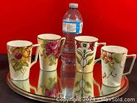 Four hand painted floral mugs arranged on a metal tray with a water bottle for scale.