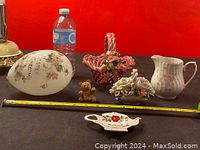 Photo of five assorted ceramic and decorative niknaks including a floral nesting egg, teddy bear figurine, pink basket, floral pitcher, and small teapot dish, with measuring tape and water bottle for scale.