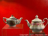 Two vintage decorative porcelain teapots displayed against a red background. One is squareish with a classical painting and purple floral accents, the other is rounded with pink roses and gold trim.