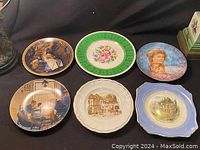 Photo of six decorative plates with various floral, portrait, and historical scene designs on a black cloth background