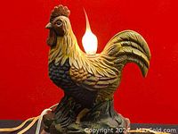 Side view of the ceramic rooster lamp with light bulb lit and cord visible.