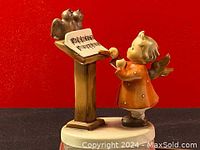 Front view of the Goebel figurine showing the angel in orange dress with wings, music stand with sheet music and two birds on top, against red background.