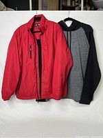 Red fleece-lined CX2 jacket and black/grey ATC hooded sweatshirt hanging side by side on a white backdrop