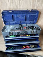 Open toolbox showing 3 drawers with tools including adjustable square, measuring tapes, screwdrivers, and pliers.