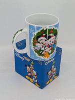 Mug on top of original Disney box showing front and side Christmas design with Mickey, Minnie, and Goofy inside wreath on light blue background, handle visible with holly decoration.