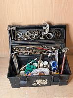 Image showing the open black plastic toolbox packed with various plumbing tools including pipe cutters, wrenches, brass fittings, pipe seats, welding torch, Teflon tapes, and assorted small hardware parts. Items show used condition with some dirt and wear.