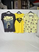 Three t-shirts hanging side by side against a white backdrop showing front designs: Jaws graphic on dark gray, yellow Superman logo, yellow butterfly print V-neck.