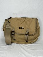 Front view of tan U.S. military style musette bag with U.S. stencil on flap and adjustable strap.