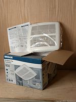 Photo of the Broan ventilation fan white grill on top of the product box, with instruction leaflet beside it.